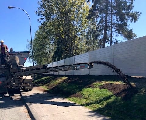 Machine slinging topsoil by highway noise barrier wall